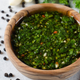 A bowl of vibrantly green chimichurri sauce in a wood bowl on a white marble counter with chili peppers, parsley, and peppercorns